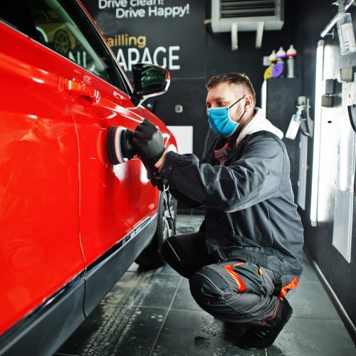 Car detailing concept. Man in face mask with orbital polisher in repair shop polishing orange suv car.